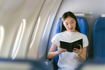 Young Asian woman traveler sitting near window and looking out window holding notebook on airplane,...