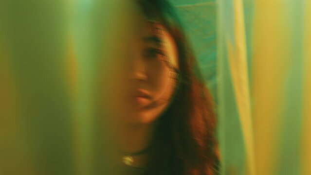 Young pretty Asian woman with hair braids looking at camera from behind curtains in studio with colorful neon light. Video portrait