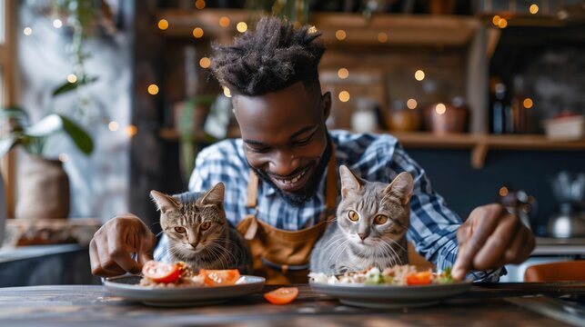 Smiling African American man preparing meals for his two cats at a modern kitchen table, surrounded by cozy, bohemian decor