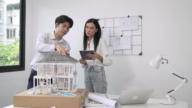 Beautiful young caucasian project manager brief asian engineer by using tablet while engineer measuring house model on meeting table with blueprint, architectural equipment scatter around. Immaculate
