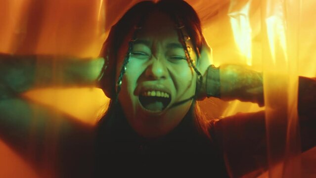 Young emotionally unstable woman covering her ears and shouting with eyes squeezed in dark studio with plastic wrap curtains and colorful illumination. Zoom away