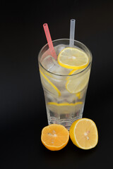 Refreshing lemonade in a tall faceted glass with ice, straws and lemon slices on a black background.