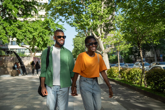 Cheerful carefree african american couple walks with hold hands in sidewalk downtown guy makes girlfriend laugh by fun storytelling. Stylish serene black young woman enjoy stroll with happy boyfriend.