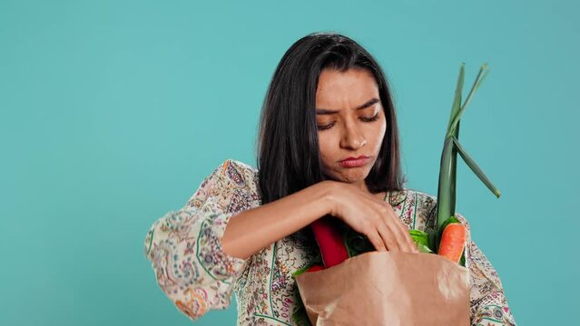 Upset woman frowning after forgetting to buy everything, looking inside paper bag with vegetables. Conscious living person sulking after realizing some groceries are missing, camera B