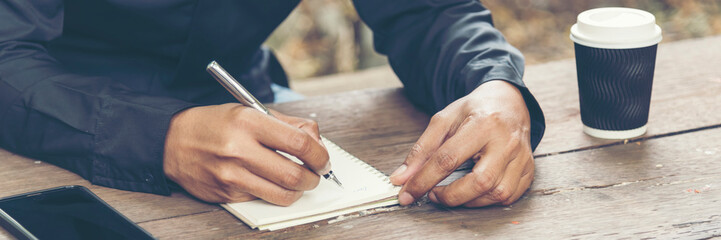 Man hands writing notebook diary with coffee cup and smartphone on wood desk. Close up man hands...