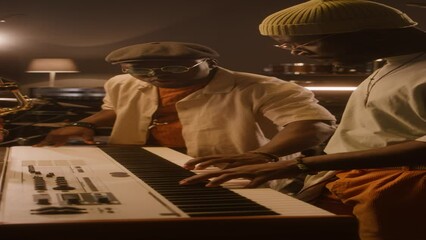 Medium vertical of two black male musicians standing at synthesizer in recording studio, playing chords on keyboards, while composing song for new album