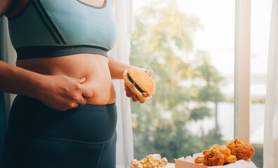 Portrait of obese woman looks sad while pinching her belly fat surrounded by junk foods. Diet fail...