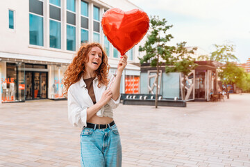 cheerful redhead woman with heart-shaped balloon walking around city 