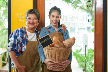 Asian Elderly mother and daughter, in aprons, stand in a bakery; older welcomes, younger showcases...