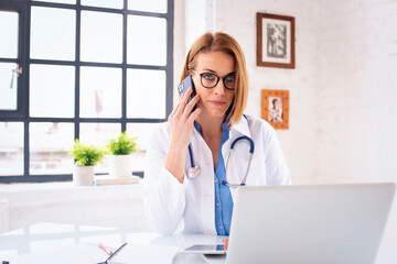 Female doctor sitting at doctor's office and using laptop and smartphone