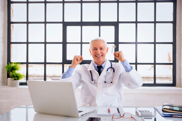 Happy male doctor sitting in doctor's room and using laptop