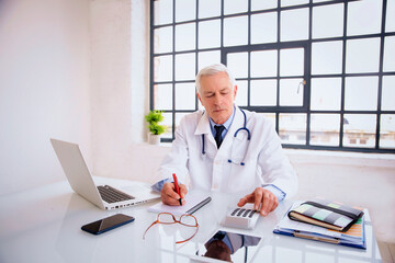 Elderly male doctor sitting in doctor's room and using laptop