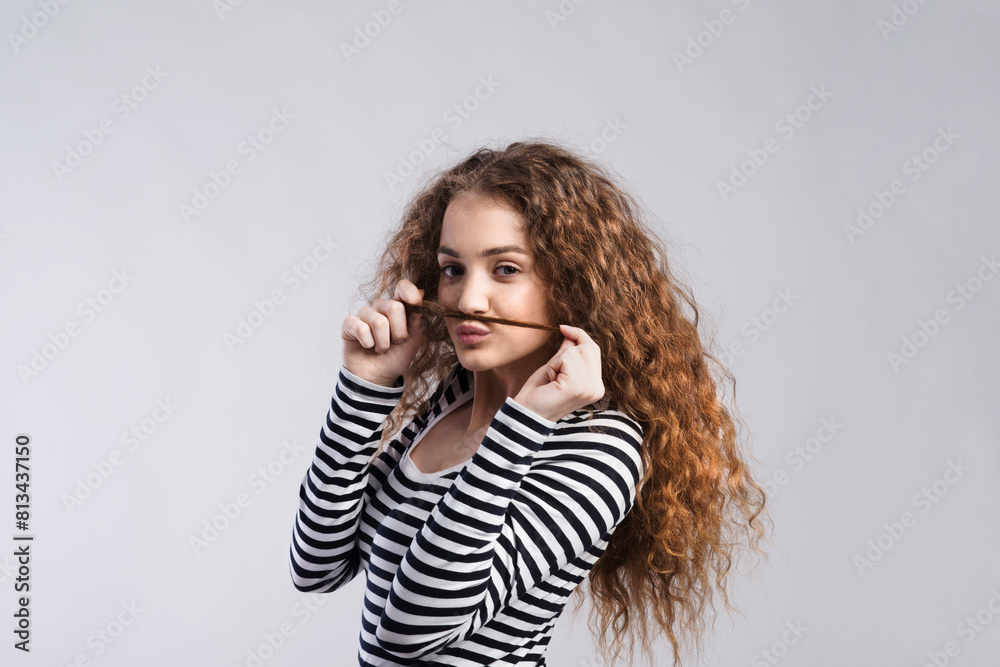 Wall mural Portrait of a gorgeous teenage girl with curly hair, holding lock of hair as a moustache. Studio shot, white background with copy space