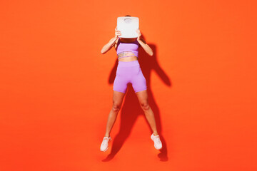 Full body young fitness trainer instructor woman sportsman wears top shorts purple clothes train in home gym jump high hold scales isolated on plain orange background. Workout sport fit abs concept.