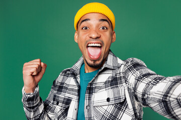 Close up young man of African American ethnicity wear shirt blue t-shirt yellow hat doing selfie shot pov on mobile cell phone do winner gesture isolated on plain green background. Lifestyle concept.