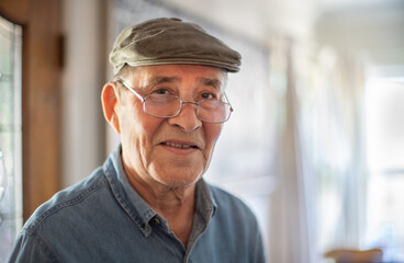 Portrait of happy smiling senior man wearing fashionable flat cap and glasses at home, closeup. Shallow DOF, copyspace.