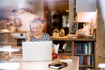 Older woman working from homeoffice, typing on laptop, making phone calls. The retiree earning...