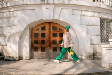 A woman in a green suit and green pants is walking down a street with a bouquet of yellow flowers...