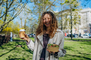Radiant young woman laughing while holding a coffee cup and a salad bowl in a vibrant city park, enjoying a sunny day and a casual meal outdoors.