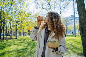 Young woman with curly hair drinks coffee and holds salad bowl while standing in sunny park, healthy lifestyle amidst lush greenery. Office employee spends her lunch break in city park.