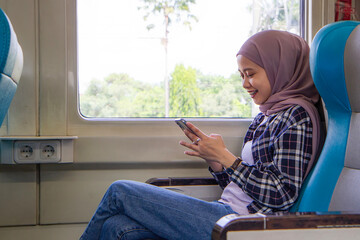 happy asian muslim woman holding her mobile phone while sitting beside big window train wagon. traveling concept