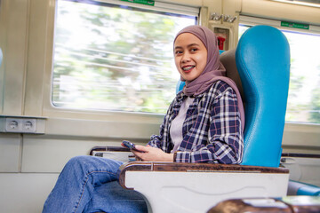 happy asian muslim woman holding her mobile phone while sitting beside big window train wagon. traveling concept