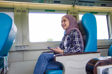 happy asian muslim woman holding her mobile phone while sitting beside big window train wagon. traveling concept