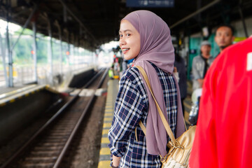 happy asian muslim woman standing on train station platform waiting for the train. traveling concept