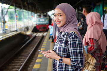 happy asian muslim woman holding phone standing on train station platform waiting for the train....