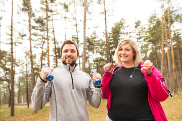 An overweight woman exercising outdoors with friend, fitness coach. Holding dumbbells.