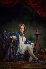 Portrait of elderly man dressed in elaborate baroque-style attire, looks as aristocratic person against vintage studio background. Concept of comparisons of eras, fusion of modernity and history. Ad