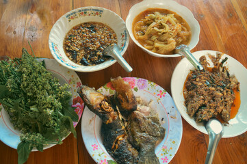 Overhead view of an authentic traditional thai meal of grilled catfish or pla duk yang, sour bamboo soup or Keng No Mai, tamarind dipping sauce and neem tips on a wooden table