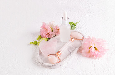 Rose quartz roller massager, gua sha scraper, massage and care oil in a matte bottle on white ceramic tray. Top view. Lifting effect.