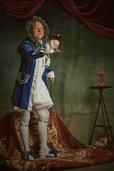 Elderly man wearing Baroque-style attire toasting and raising hand with glass of red wine against...