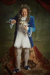 Elderly man dressed in richly detailed baroque costume, holding and admiring fish against vintage...