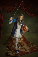 Elderly man wearing in baroque costume holding sword, sitting on playful horse-toy against vintage...