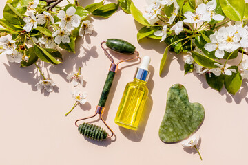 roller massager, gua sha scraper made of natural stone, oil in a bottle with a pipette among the flowering branches of the fruit tree. Natural care.