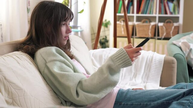 Child, teenage is sitting on the couch at home, using the remote control to switch channels on the TV.