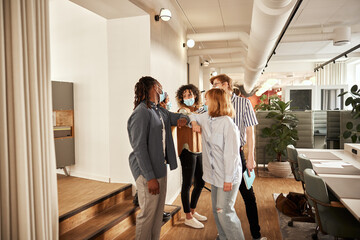 Diverse business people greet each other with elbows bumps, they are all wearing a blue face mask, in an open office space.