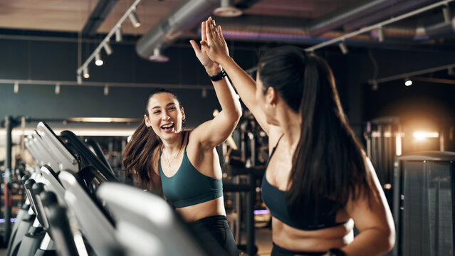 Two ecstatic fit young women in sportswear laughing and high-fiving each other after a cardio workout session on stationary bikes in a gym