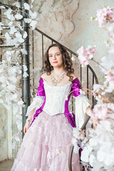 Beautiful woman in fantasy white and purple rococo style medieval dress standing on the stairway with white flowers