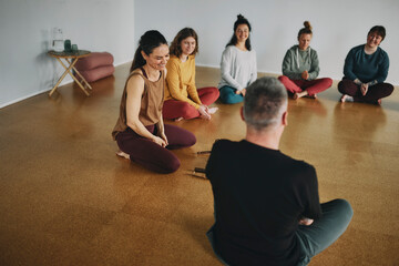 Female yoga instructor talking and laughing with a group of students before a meditation class together on the floor of her studio