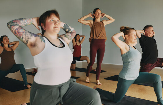 Smiling female yoga teacher leading a group of students through their poses during a class in her studio. The focus is on a young woman with tattoos and hair under her arms