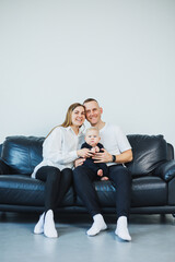 A young married couple with a small child is photographed sitting on a sofa on a white background. Caring parents look at their baby while holding it in their arms.