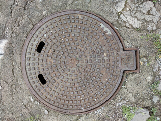 The image shows a round, metal manhole cover with a grid pattern on its surface. The cover is set...