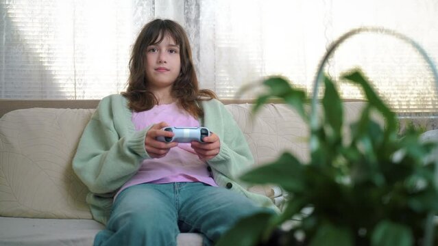 11 years old child holding gaming console and playing on a sofa. Portrait of pre-teenage girl with video game remote control playing a video game, holding joystick, smiling. Children's Leisure And