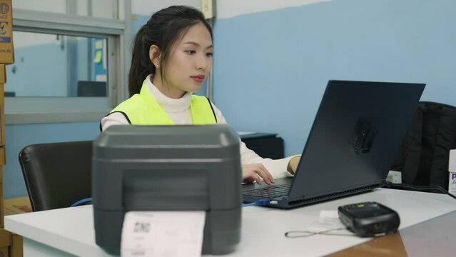 Beautiful Asian woman uses a laptop or notebook to print bar code stickers on a bar code printer. Selective focus at sticker barcode. Selective focus at barcode printer.