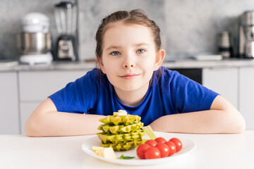 Portrait of a happy little girl having breakfast in the kitchen at home. Healthy breakfast of waffles with spinach, cheese and tomatoes. Selected focus.