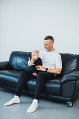 Young adult surprised man wearing casual style maroon t shirt holding baby girl or boy in arms and using mobile phone while sitting on sofa in living room, get shocking news from email.