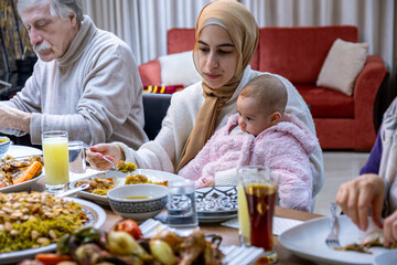 A tender portrait captures a mother seated at the family table, lovingly caring for her baby while they share a meal together. The bond between them shines through as they create cherished memories 